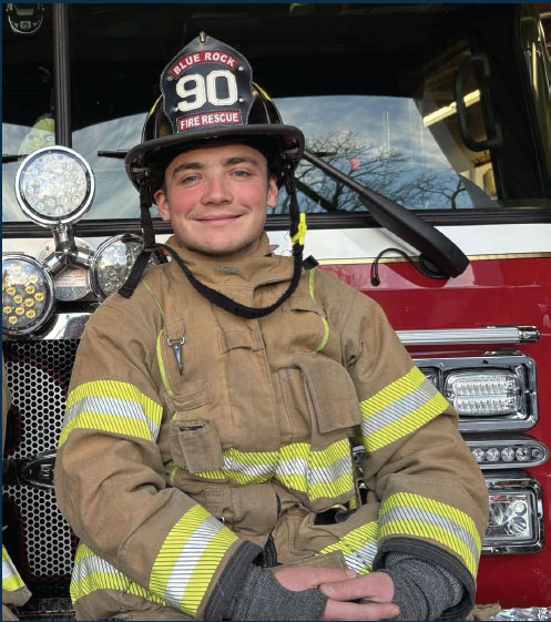 Jake Speers leaning against a fire truck in fire gear